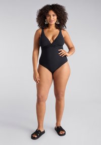 Svart baddräkt med djup V-ringning och knutdetalj vid midjan, med texturerade utsmyckningar på bröstområdet.