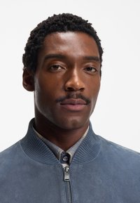 Giacca bomber in camoscio azzurro chiaro con colletto a coste e chiusura con zip. Presenta una finitura testurizzata e una camicia stratificata sotto.