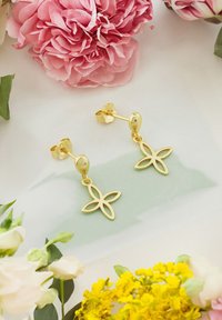 Guldhängande örhängen med en fyrblommig design. Slät yta med glänsande finish, i kombination med blomsterelement i bakgrunden.