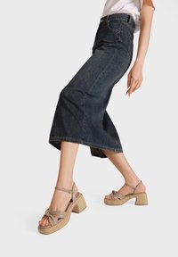 Sandali platform color marrone chiaro con dettaglio a nodo, texture intrecciata sui tacchi, abbinati a una gonna in denim scuro con silhouette svasata.