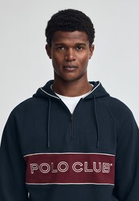 Felpa navy con collo a zip a un quarto, caratterizzata da una striscia orizzontale bordeaux con "POLO CLUB" ricamato in bianco. Materiale in cotone, vestibilità comoda.