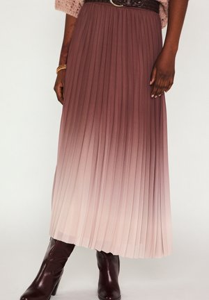 Femme portant une jupe plissée ombrée du marron au rose, des bottes à talons brun foncé, une ceinture marron et un pull tricoté rose, debout devant un fond blanc.