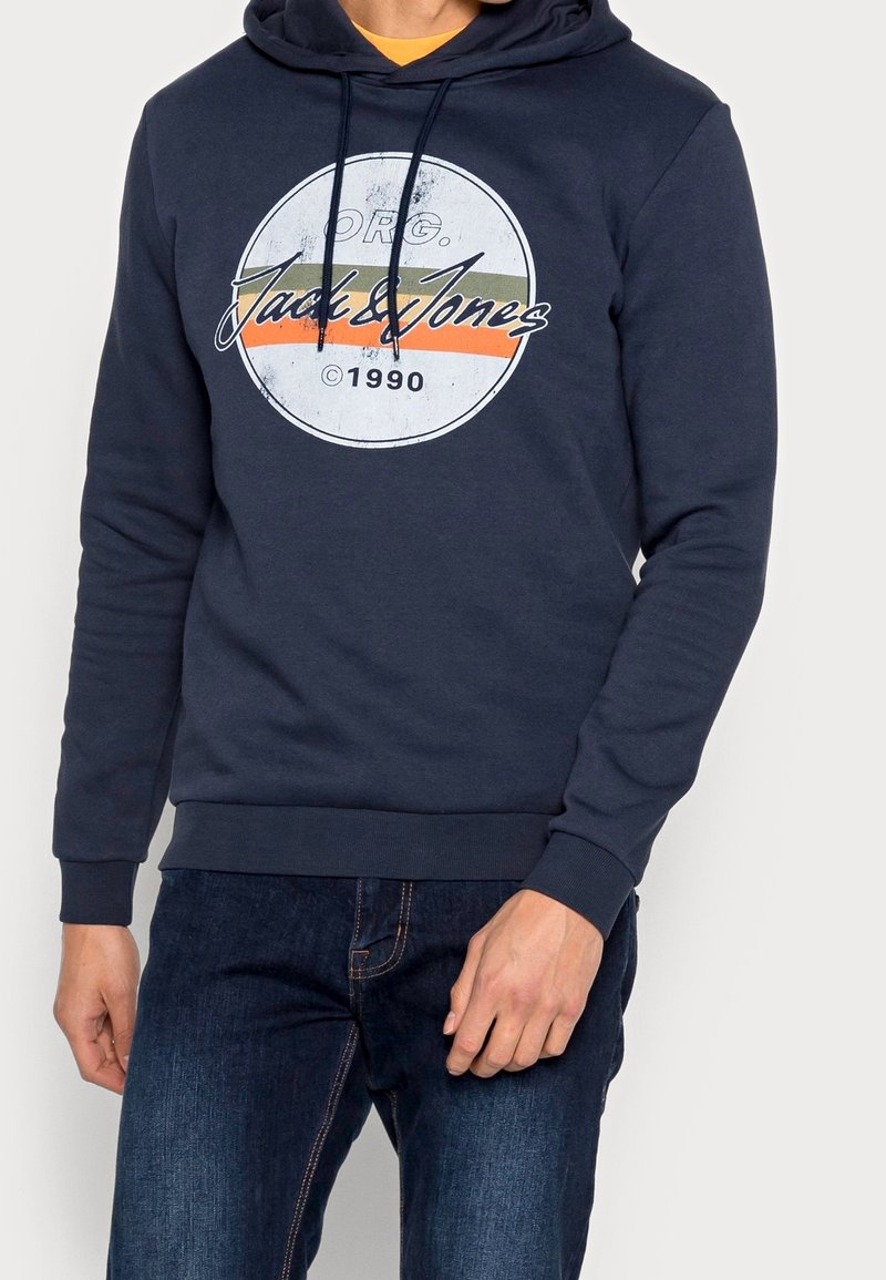 Homme portant un sweat à capuche bleu marine avec un logo blanc rond et le texte « Jack & Jones © 1990 » sur des bandes orange et vertes, associé à un jean bleu foncé.
