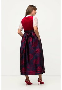 Frau in einem roten Samt-Top mit Spitzenärmeln, einem marineblauen und roten Blumenkleid und roten, hochhackigen Slingbacks, die seitlich steht.