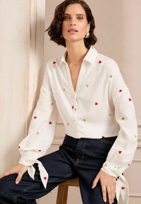 Camicia bianca con ricamo a forma di cuore rosso e oro, maniche lunghe e puff con dettaglio a cravatta, abbinata a pantaloni in denim scuro.