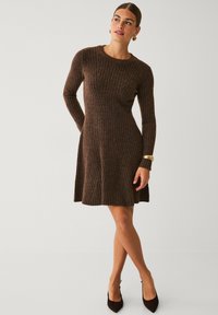 Frau in einem braunen gerippten, langärmligen Kleid mit goldenen Armbändern und Ohrringen, die mit übergeschlagenen Beinen posiert und leicht zur Seite schaut.