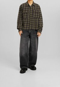 Camisa de franela a cuadros verde y negra con dos bolsillos en el pecho, combinada con jeans de pierna ancha negros desgastados y botas negras gruesas.