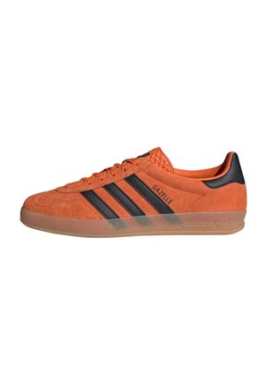 Orange ruskindsneakers med sorte striber, rund tå og gennemsigtig gummisål. Har "GAZELLE" branding på siden.