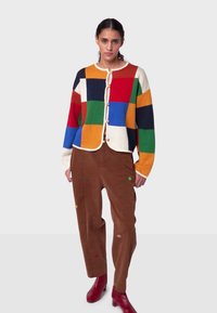 Monivärinen tilkkutäkkineuletakki, jossa on kermaiset reunukset ja napitus, päällä ruskeat puuvillafarkut, joissa on hillittyjä kirjailuja, ja punaiset saappaat.