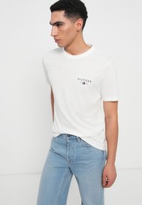 Tricou alb din bumbac cu un mic logo negru "HILFIGER 1985" pe partea stângă a pieptului, purtat cu blugi din denim bleu deschis.