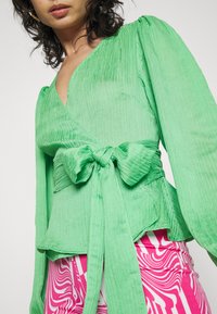 Blusa verde a coste con maniche lunghe e scollatura a V, dotata di cintura da annodare in vita. Abbinata a pantaloni rosa a fantasia con motivi a spirale.