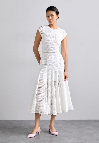 Robe midi blanche à motifs avec des manches courtes, un corsage texturé, une jupe évasée et une fine ceinture. Associée à des mules à talons rose pâle.