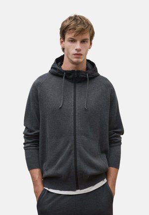 Joven con cabello castaño claro que lleva una sudadera con capucha y cremallera de color gris oscuro con capucha negra y las manos en los bolsillos.