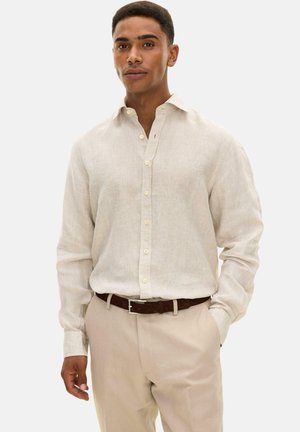 Mann trägt ein beiges, langärmliges Hemd mit Knöpfen, das in eine hellbraune Hose mit einem dunkelbraunen Gürtel gesteckt ist, steht mit einer Hand in der Tasche.