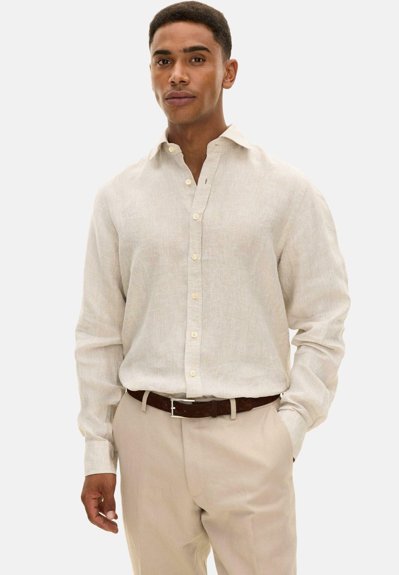 Mann trägt ein beiges, langärmliges Hemd mit Knöpfen, das in eine hellbraune Hose mit einem dunkelbraunen Gürtel gesteckt ist, steht mit einer Hand in der Tasche.