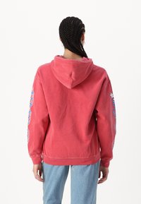 Rød hettegenser med snor og logo-detalj på venstre erme. Myk stoff og avslappet passform, kombinert med lyseblå denimjeans.