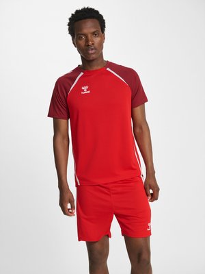 Man standing facing forward wearing a red sports outfit with maroon sleeves and white accents, looking straight ahead with a neutral expression.