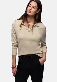 Femme aux longs cheveux bruns portant un haut beige à manches longues et un pantalon noir, une main dans la poche, regardant légèrement sur le côté.