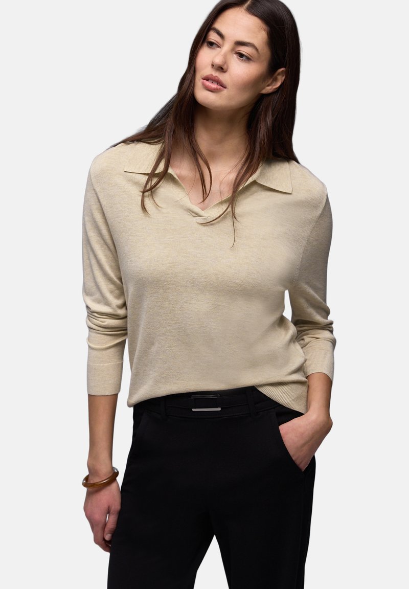 Femme aux longs cheveux bruns portant un haut beige à manches longues et un pantalon noir, une main dans la poche, regardant légèrement sur le côté.