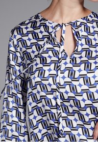 Blusa a maniche lunghe di colore azzurro chiaro con un motivo geometrico di cerchi bianchi e neri, caratterizzata da un collo con chiave e lacci.