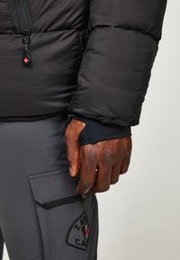 Schwarze Steppjacke mit einer strukturierten Oberfläche, ausgestattet mit einem Reißverschluss und Daumenloch-Bündchen. Graue Cargohosen mit Tasche und Logodetail.