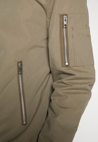 Kurtka oliwkowo-zielona o gładkiej, teksturowanej powierzchni. Posiada zapinane na zamek kieszenie na rękawie oraz ukryty zamek z przodu.