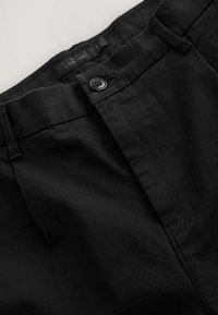 Pantalon en coton noir à texture lisse, doté d'une fermeture à boutons, de passants pour ceinture et d'une étiquette sur la ceinture.
