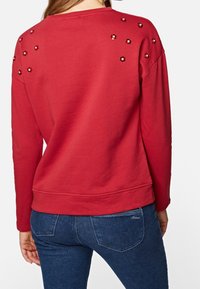 Roter Sweatshirt mit langen Ärmeln, das mit Pailletten-Blumenakzenten auf der Rückseite verziert ist. Weicher Stoff, abgerundeter Saum und lockere Passform. Kombiniert mit blauen Jeans.