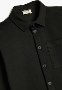 Camicia nera con bottoni, colletto strutturato, tessuto testurizzato, sei bottoni abbinati e taschino a sinistra sul petto. Etichetta del marchio Cotton Kids visibile.
