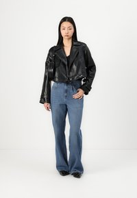 Veste en cuir noire courte avec grands revers, associée à un jean bleu évasé taille haute. Le mannequin tient un petit sac et porte des chaussures noires.