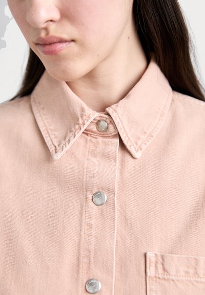 Šviesiai rožinė džinsinė marškiniai su smailiu apykakle, sidabriniais užsegimais ir priekiu kišenė. Audinys lygus, matomi siuvimo detalės.