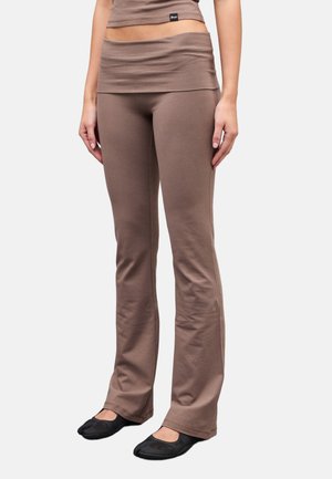 Mujer con pantalones acampanados ajustados de color marrón y zapatos negros sin cordones, de pie frente a un fondo claro y liso.