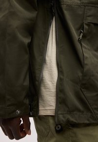 Delvis bilde av en person som har på seg en oppglidset mørkegrønn jakke over en lys beige skjorte og matchende grønne bukser, hånden avslappet ved siden.