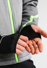 Veste grise avec une fermeture éclair et des accents verts. Dotée de poignets noirs, d'une texture brossée et d'une coupe ergonomique autour des mains.