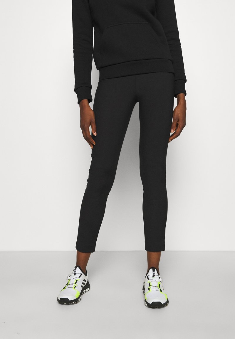 Personne portant un pantalon noir ajusté, un sweat-shirt noir et des chaussures de sport blanches avec des accents jaune néon, debout devant un fond clair uni.