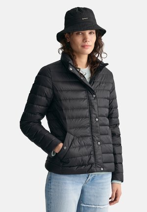 Doudoune noire avec col ajusté, design matelassé en diagonale, boutons-pression à l'avant et poches latérales, portée avec un jean bleu clair.