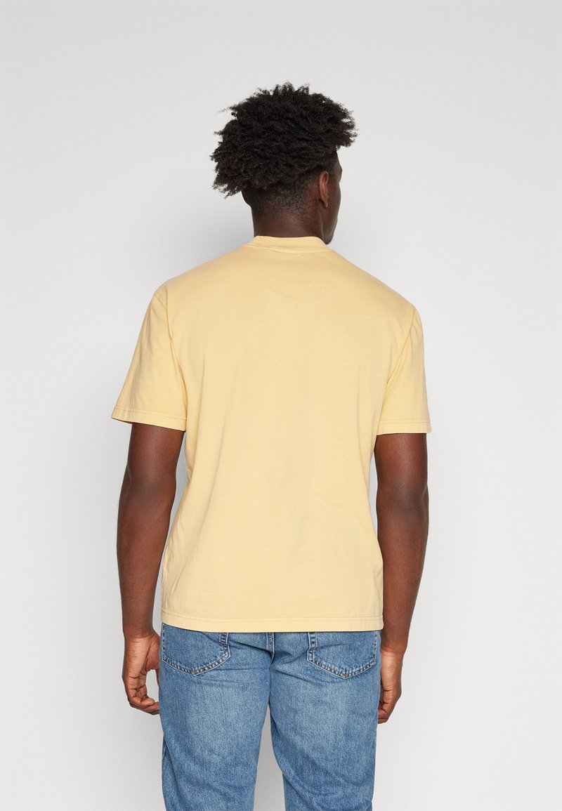 Żółta krótki rękawek t-shirt z okrągłym dekoltem, gładką teksturą i gładkim tyłem. Noszony z niebieskimi dżinsami, z delikatnymi detalami na tylnych kieszeniach.