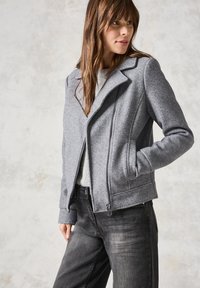 Veste en laine grise avec une fermeture éclair diagonale, de larges revers et deux poches latérales. Portée sur un pull léger et associée à un jean foncé.