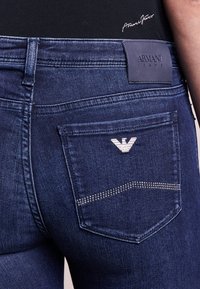 Jeans in denim blu scuro con vestibilità slim, caratterizzati da un'etichetta in pelle sulla cintura, dettagli nelle tasche e un logo argentato sulla tasca posteriore.