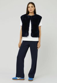 Gilet en fausse fourrure marine avec un col rond et des boutons-pression à l'avant, porté sur un t-shirt blanc. Associez-le à un pantalon large marine et des baskets bleues.