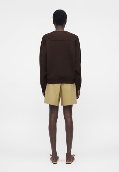 Άτομο με κοντά σγουρά μαλλιά που φοράει σκούρο καφέ μακρυμάνικο μπλουζάκι, μπεζ σορτς μέχρι τα γόνατα και καφέ σανδάλια, στέκεται με την πλάτη προς τα εμπρός.