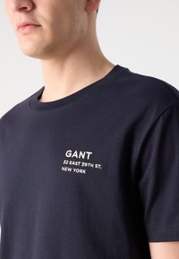 Tamsiai mėlynas medvilninis marškinėliai su apvaliu kaklu, su baltais siuviniais: "GANT 32 EAST 39TH ST. NEW YORK" kairiajame krūtinės kampe.