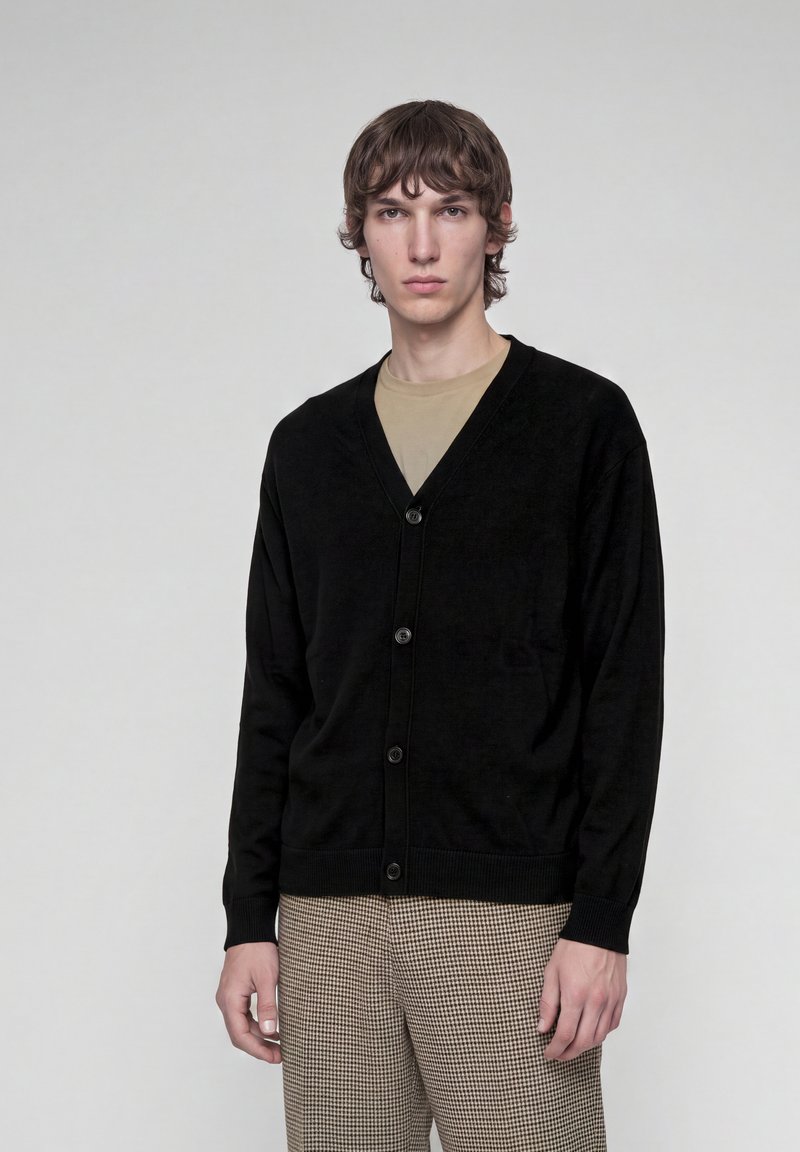 Jeune homme portant un cardigan noir boutonné sur une chemise beige et un pantalon marron à carreaux, debout devant un fond clair uni.