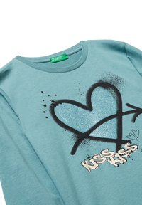 Světle modrá košile s dlouhým rukávem, na které je třpytivé srdce a černý design šipky, s efekty sprejového malování a textem "KISS KISS" v krémových písmenech.