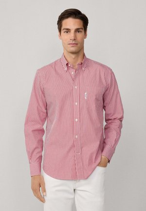 Homme portant une chemise à boutons rouge et blanche à rayures verticales avec poche, associée à un pantalon blanc, se tenant devant un fond gris clair.