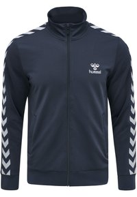 Marine sportjack met hoge kraag, ritssluiting en twee zijzakken. Heeft witte chevronstrepen langs de mouwen en een logo op de borst.