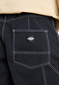 Pantalons en tissu noir avec une poche arrière dotée d'une étiquette logo rectangulaire, des coutures blanches contrastantes et une coupe ajustée.