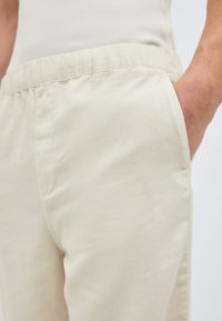 Άτομο που φοράει ανοιχτό μπεζ παντελόνι με ελαστική μέση, με το χέρι στην πλαϊνή τσέπη και λευκό ριμπ τοπ μαζεμένο μέσα.