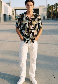 Young man wearing a black tropical print short-sleeve shirt, white pants, and white sneakers, standing outdoors on a sunlit concrete surface.