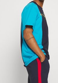 Tričko s barevným blokováním v tyrkysové a námořnické barvě, s kulatým výstřihem, krátkými rukávy, s žlutým logem na detailu. Spárováno s námořnickými kalhotami.